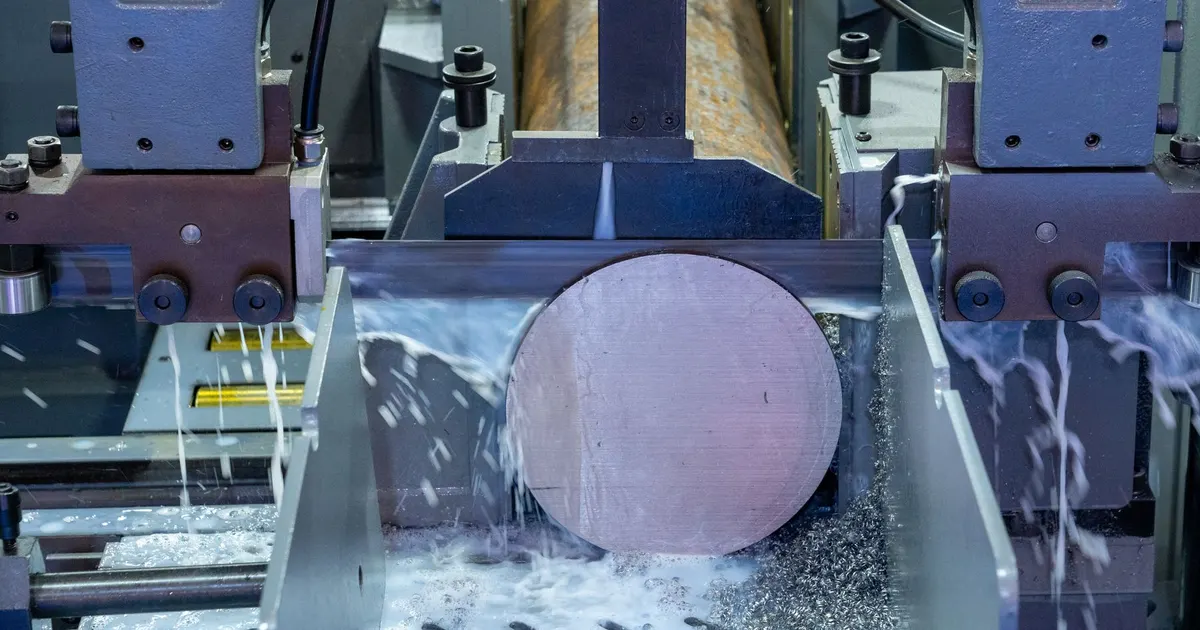 Band saw cutting through a round metal bar with coolant flowing over the blade, a typical first step when working from bar stock