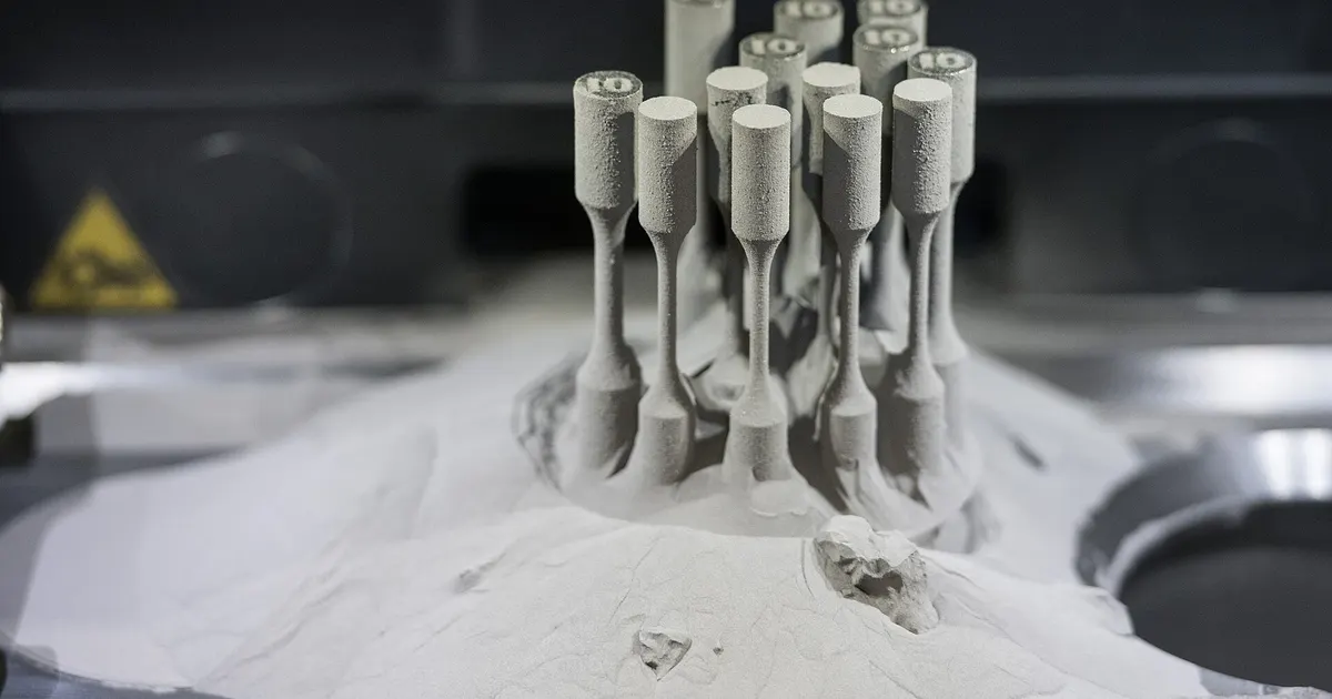 Small metal test bars standing on a build plate in loose powder inside a laser powder bed fusion machine, the starting point for machining additive Inconel 718 parts